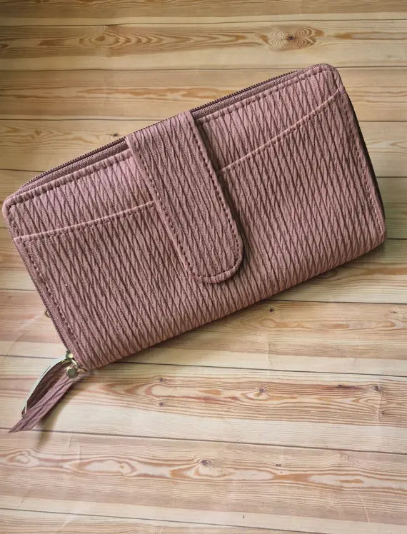 A dusty rose pink zip-around wallet with a textured, vertically ribbed fabric and a matching tassel zipper pull, displayed diagonally on a light-colored wooden plank surface. Main image
